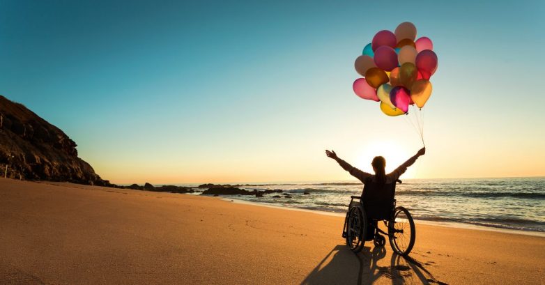 Person in wheelchair holding colorful balloons at sunset beach